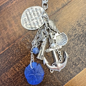 May include: A silver-toned keychain with nautical charms, including an anchor, lighthouse, seahorse, and a blue glass sand dollar. A circular charm has the text: "may always have a shell in your pocket & sand between your toes."
