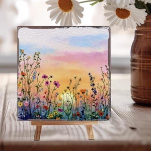 May include: A square watercolor painting of a sunset over a field of wildflowers, displayed on a small wooden easel. The painting features a vibrant sky with hues of pink, orange, and blue. The wildflowers are in shades of purple, yellow, and red.