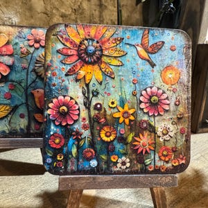 May include: Square wooden art piece featuring a vibrant floral design with a large sunflower, hummingbirds, and various colorful flowers. The artwork is displayed on a small wooden easel, showcasing a rustic, artistic style.
