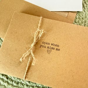 May include: A brown kraft paper envelope tied with a piece of natural jute twine. The front of the envelope features the black stamped text "open when you miss me" followed by a small hand-drawn heart symbol. A white card is visible inside.