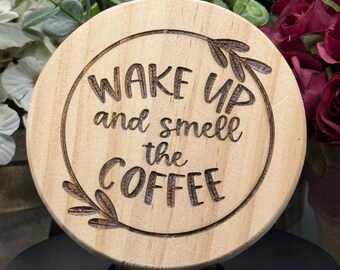 May include: A round, wooden coaster with the phrase "WAKE UP and smell the COFFEE" etched into the surface. The coaster has a light wood grain and a decorative leaf design. The coaster is a coffee-themed item.