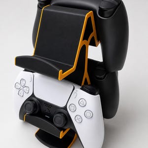 May include: A black and orange plastic controller stand holding two gaming controllers. One controller is white with black accents and the other is solid black. The stand features a tiered design with a central support structure and a base.