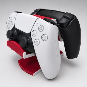 May include: A red plastic dual controller stand holding a white and black wireless game controller in the front and a solid black wireless game controller in the back. The stand features a curved design to support the controllers securely.