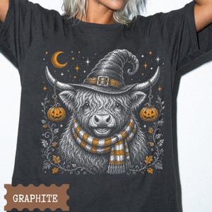 May include: A graphite gray t-shirt featuring a detailed graphic of a Highland cow wearing a witch hat and a striped scarf. The cow is flanked by two hanging jack-o'-lanterns, with a crescent moon, stars, and autumn leaves in the background.