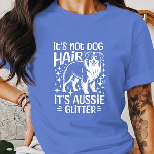 May include: A blue t-shirt with white text that reads "it's not dog hair it's Aussie glitter" and a white silhouette of an Australian Shepherd dog.