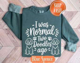 Può includere: Felpa a maniche lunghe blu abete con la scritta bianca "I was normal two doodles ago". Include illustrazioni di cani barboncino e impronte di zampe. La parte inferiore della maglietta riporta la scritta "Blue Spruce".