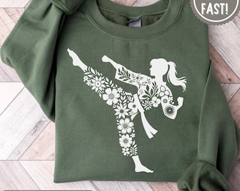 May include: A military green crewneck sweatshirt featuring a white graphic print of a person in a martial arts pose. The silhouette of the figure is filled with a detailed floral pattern. The sweatshirt has long sleeves and a ribbed collar.