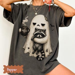 May include: A charcoal gray t-shirt featuring a graphic of a ghost holding a mug with the text MEH and embracing a raccoon. The ghost has a stitched patch on its head, and small bats and a tiny ghost icon are printed around the central illustration.