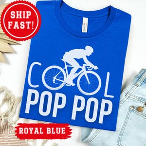 Puede incluir: Camiseta azul real con un gráfico blanco de un ciclista en bicicleta y las palabras "COOL POP POP". La camiseta está hecha de un material suave.