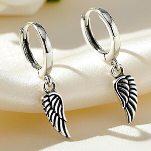 May include: A pair of silver hoop earrings with dangling wing charms. The earrings have a polished finish and a hinged closure. The wing charms are detailed with etched lines, creating a feather-like appearance. The earrings are displayed on a white satin surface.