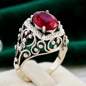 May include: A silver ring featuring a large, oval-cut, deep red gemstone set in a prong setting. The band and gallery are crafted with intricate, openwork filigree metal patterns, including swirling scrolls and small spherical bead accents.