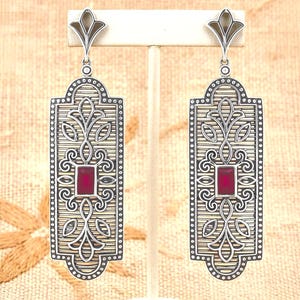 May include: A pair of long, rectangular silver-toned earrings featuring an intricate filigree pattern with a central, vertical, deep red rectangular gemstone. The earrings have a pointed, fan-like top element and a textured, linear background.