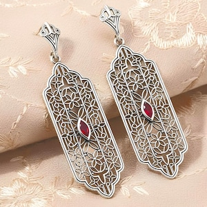 May include: A pair of silver-toned filigree earrings. Each earring features an elongated rectangular shape with a floral design and a central, oval-shaped red gemstone. The earrings have a decorative top and a hook closure.