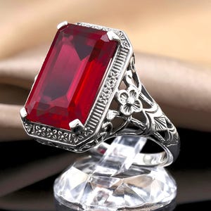 May include: A silver ring featuring a large, rectangular, deep red gemstone set in a frame with intricate metalwork. The band displays detailed floral and geometric engravings along the sides, supporting the prominent, faceted red stone.