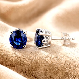 May include: A pair of stud earrings featuring oval-cut, deep blue gemstones set in silver-toned metal. Each earring has a four-prong setting with a decorative, openwork gallery design and a standard post back with a matching metal butterfly clutch.