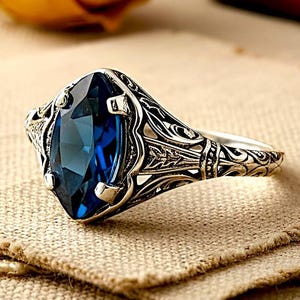 May include: A silver ring with an oval, deep blue gemstone. The ring features intricate filigree detailing around the stone and along the band. The gemstone is set in a four-prong setting. The ring is displayed on a tan fabric.