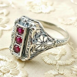 May include: A silver ring with a rectangular face featuring three round, red gemstones. The ring has an ornate, filigree design around the gemstones and along the band, showcasing intricate details and a vintage aesthetic.