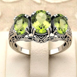 May include: A silver ring featuring three oval-cut, light green gemstones. The ring has an ornate, filigree design on the band. The gemstones are set in a prong setting, and the ring is displayed on a white surface.