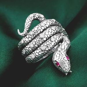 May include: A silver snake ring with a coiled design. The ring features small, clear gemstones and red gemstones for the snake's eyes. The ring is set against a dark green background.