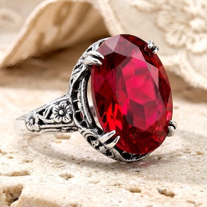 May include: A silver-toned ring featuring a large, oval-cut, deep red gemstone held by four prongs. The band and setting are crafted with intricate, raised floral and vine-like metalwork details that extend along the sides of the ring.