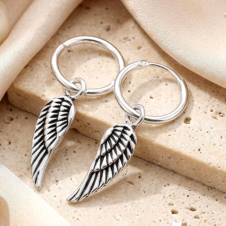 May include: A pair of silver hoop earrings with dangling wing charms. The wings have detailed feather designs with black accents. The earrings are displayed on a textured, light-colored surface with a soft, draped fabric in the background.