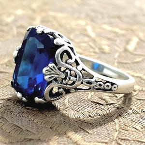 May include: A silver-toned ring featuring a large, rectangular, deep blue faceted gemstone set within an intricate, openwork metal band with scrolling, filigree-style patterns and darkened recessed areas for contrast.