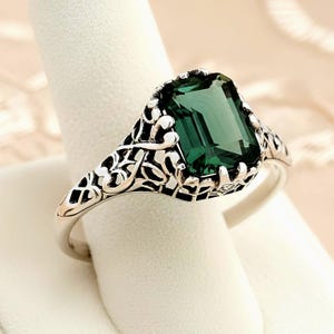 May include: A silver ring featuring an emerald-cut, deep green gemstone. The ring has an ornate, filigree design on the band and around the gemstone setting. The ring is displayed on a white surface, showcasing its intricate details and elegant style.