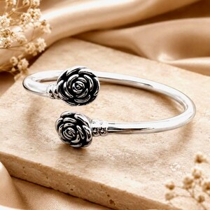 May include: A silver-toned open cuff bracelet featuring two detailed rose-shaped ornaments at each end. The metal has a polished finish with darkened recessed areas within the petals to highlight the floral design of the two roses.