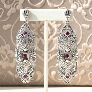 May include: A pair of silver-toned Art Deco style earrings. Each earring features an elongated, openwork design with floral and geometric patterns. The earrings are adorned with small, square, and round red gemstones and small white pearls.