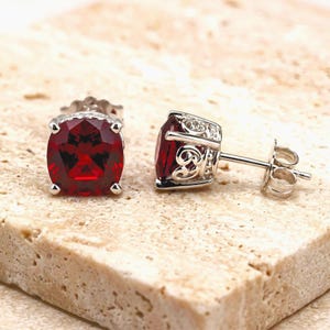 May include: A pair of stud earrings featuring cushion-cut deep red gemstones set in silver-toned metal. The settings display an intricate openwork scroll design along the sides, and each earring includes a standard post and butterfly back closure.