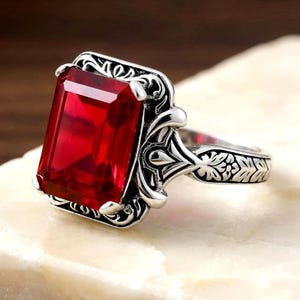 May include: A silver ring featuring a large, rectangular, faceted red gemstone set in a frame with intricate, raised scrollwork and floral engravings along the band and the setting, showcasing a polished metallic finish and deep red color.