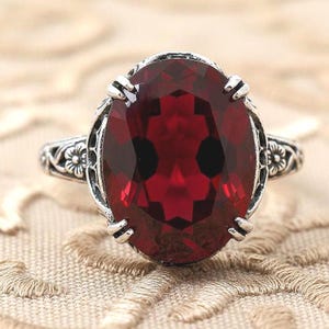 May include: A silver ring with an oval, deep red gemstone. The gemstone is set in a detailed, ornate silver band with floral accents on the sides. The ring is displayed on a textured, neutral-toned surface.