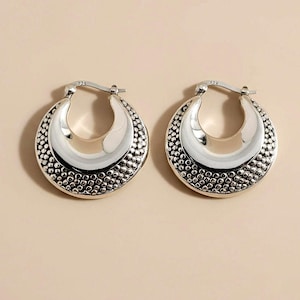 May include: A pair of silver-toned hoop earrings. Each earring has a polished, crescent-shaped inner section and a textured, dotted outer edge. The earrings are displayed against a neutral background.