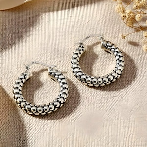 May include: A pair of silver hoop earrings. Each earring features a textured design of small, round beads. The earrings are displayed on a neutral-toned surface, with soft lighting and a hint of dried flowers in the background.