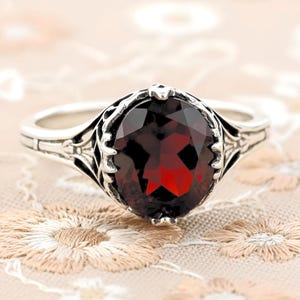 May include: A silver ring featuring a large, oval-cut, deep red garnet gemstone set in a decorative, pronged metal mounting with intricate, engraved detailing along the band, showcasing a polished finish and a dark, faceted stone center.