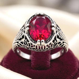 May include: A silver ring featuring a large, oval-cut, deep red gemstone set in a prong setting. The band is crafted with intricate, openwork metal detailing that extends from the central stone setting down along the sides of the ring shank.