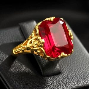 May include: A gold ring featuring a large, rectangular, faceted red gemstone set in a decorative, openwork metal band. The setting displays intricate, filigree-style metalwork surrounding the base of the vibrant, deep red stone.