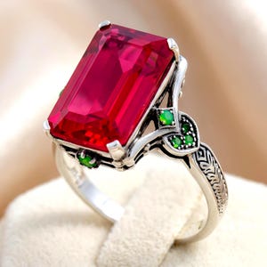 May include: A silver ring featuring a large, rectangular, faceted red gemstone as the centerpiece. The metal band is decorated with intricate engravings and accented with small, round, green stones set along the sides of the setting.