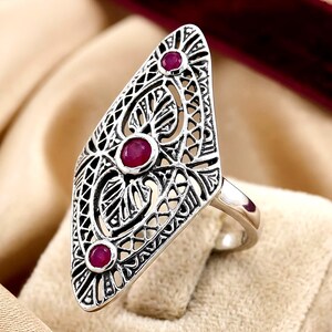 May include: A silver-toned ring featuring an elongated, diamond-shaped face with intricate openwork filigree patterns. Three small, round, deep pinkish-red gemstones are set vertically along the center of the decorative metal surface.