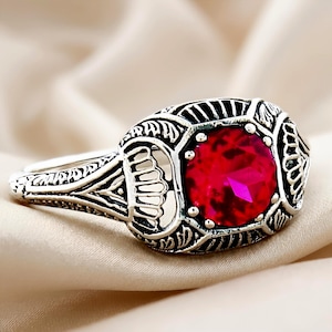 May include: A silver-toned ring featuring an oval-shaped setting with intricate, dark-oxidized filigree metalwork. A single, round, faceted bright red gemstone is securely held in the center by six small prongs, creating a high-contrast appearance.