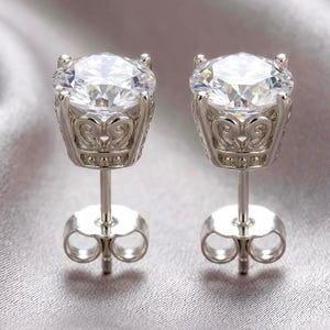 May include: A pair of silver-toned stud earrings featuring large, round, clear faceted stones. Each stone is held by a four-prong setting with an intricate, openwork scroll design along the sides and a post back with a matching silver-toned butterfly clutch.