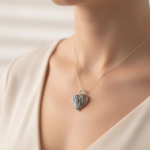 Op de afbeelding: Een zilveren hartvormige hangerketting met een gedetailleerd vleugelontwerp. De hanger hangt aan een fijne ketting. De ketting wordt gedragen door een persoon die een cr&egrave;mekleurige top draagt.