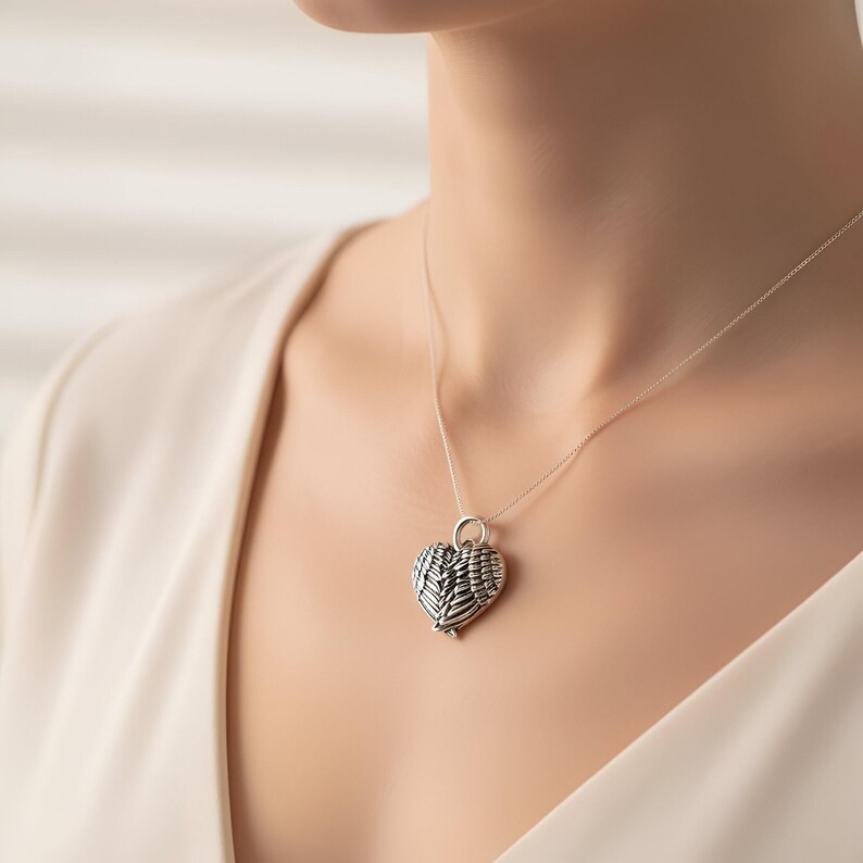 Op de afbeelding: Een zilveren hartvormige hangerketting met een gedetailleerd vleugelontwerp. De hanger hangt aan een fijne ketting. De ketting wordt gedragen door een persoon die een cr&egrave;mekleurige top draagt.