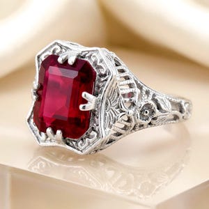 May include: A silver-toned ring featuring a large, rectangular, deep red gemstone set in a prong setting. The band and setting are crafted with intricate, openwork metal filigree patterns, including small floral motifs along the sides.