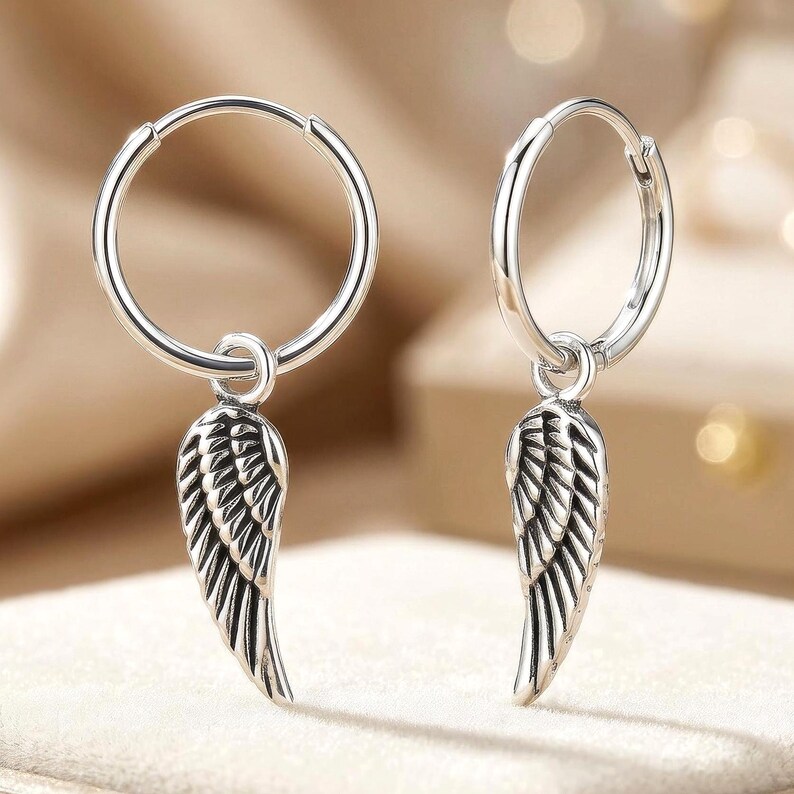 May include: A pair of silver hoop earrings, each featuring a detailed wing charm. The earrings are made of a shiny, metallic material and are designed to be worn as a fashion accessory. The wing charms have intricate feather detailing.