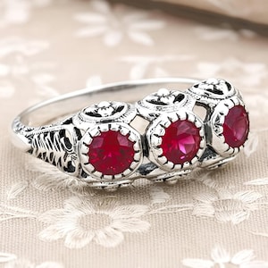 May include: A silver ring featuring three round, vibrant red gemstones. The ring has an ornate, filigree design with detailed patterns around the gemstones and along the band. The gemstones are set in a bezel setting, and the ring appears to be made of sterling silver.