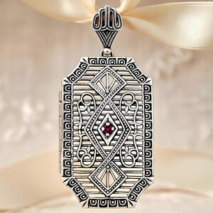 May include: Silver rectangular locket with intricate geometric patterns and a small, round, red gemstone at the center. The locket is suspended from a cream-colored ribbon, showcasing its ornate design and vintage aesthetic.