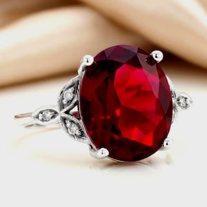 May include: A silver ring featuring a large, oval-cut deep red gemstone as the centerpiece. The band is crafted with intricate metalwork and accented by small, clear, round-cut stones embedded along the sides of the setting.