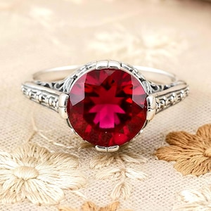 May include: A silver ring featuring a large, round, faceted deep red gemstone set in a decorative metal band with intricate, textured detailing along the sides. The setting holds the stone securely with multiple small, rounded prongs.