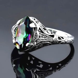 May include: A silver ring featuring a marquise-cut iridescent gemstone with a multi-colored surface. The metal band displays intricate, raised filigree patterns and scrollwork details that extend from the central setting along the sides.